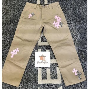 Può includere: Pantaloni color cachi con diverse toppe a forma di croce rosa e dettagli a scacchi bianchi e neri. I pantaloni hanno un classico design a cinque tasche e una chiusura con bottone e cerniera. Il testo "Best Two Shop" è visibile.