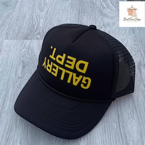 Puede incluir: Gorra de camionero negra con la inscripción "GALLERY DEPT." en amarillo. La gorra tiene una parte trasera de malla y una visera curva. Se encuentra sobre una superficie de madera gris claro.