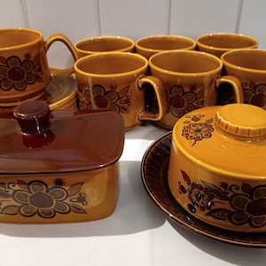 May include: A vintage ceramic tableware set in shades of brown and gold. The set includes mugs, saucers, a creamer, a covered dish, and a sugar bowl. Each piece is decorated with a floral design.
