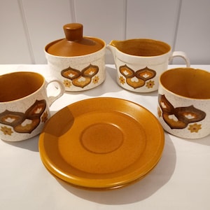 May include: A retro ceramic coffee set featuring a speckled white finish and brown and orange floral patterns. The set comprises two mugs, a lidded sugar bowl, a creamer jug, and a saucer. The design suggests a 1970s origin.
