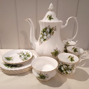 May include: A white porcelain tea set with a floral design. The set includes a teapot, cups, saucers, and a sugar bowl. The floral pattern features white flowers with green leaves and gold trim. The set is arranged on a white surface.