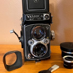 May include: A black Yashica Mat-124 twin-lens reflex camera with a lens hood, lens cap, and cleaning brush. The camera has a silver and black design with the brand name prominently displayed. The camera is on a wooden surface.