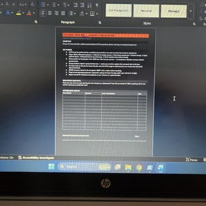 May include: A computer screen displays a document with text and tables. The document has a red and orange header. The screen shows a Microsoft Word interface with various editing tools and a search bar at the bottom.