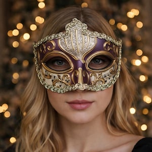 Puede incluir: Una máscara decorativa de carnaval con una combinación de colores púrpura, dorado y crema. La máscara presenta detalles ornamentados, incluyendo notas musicales y detalles brillantes. Diseñada para cubrir los ojos y la parte superior de la cara, ideal para un baile de máscaras.