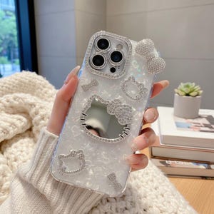 K&ouml;nnte beinhalten: Eine transparente Handyh&uuml;lle mit wei&szlig;em Perlmutt-Design, verziert mit glitzernden Strasssteinen. Die H&uuml;lle zeigt ein Hello Kitty-Design, einschlie&szlig;lich einer Schleife und dem Gesicht der Figur. Das Kameraobjektiv ist von einem Strasssteinrand umgeben.
