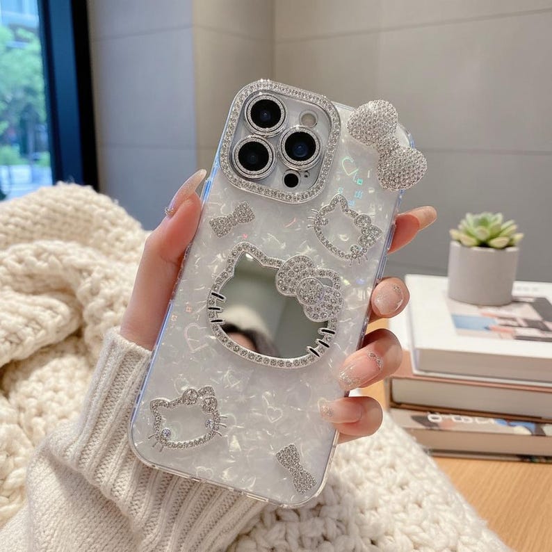 K&ouml;nnte beinhalten: Eine transparente Handyh&uuml;lle mit wei&szlig;em Perlmutt-Design, verziert mit glitzernden Strasssteinen. Die H&uuml;lle zeigt ein Hello Kitty-Design, einschlie&szlig;lich einer Schleife und dem Gesicht der Figur. Das Kameraobjektiv ist von einem Strasssteinrand umgeben.