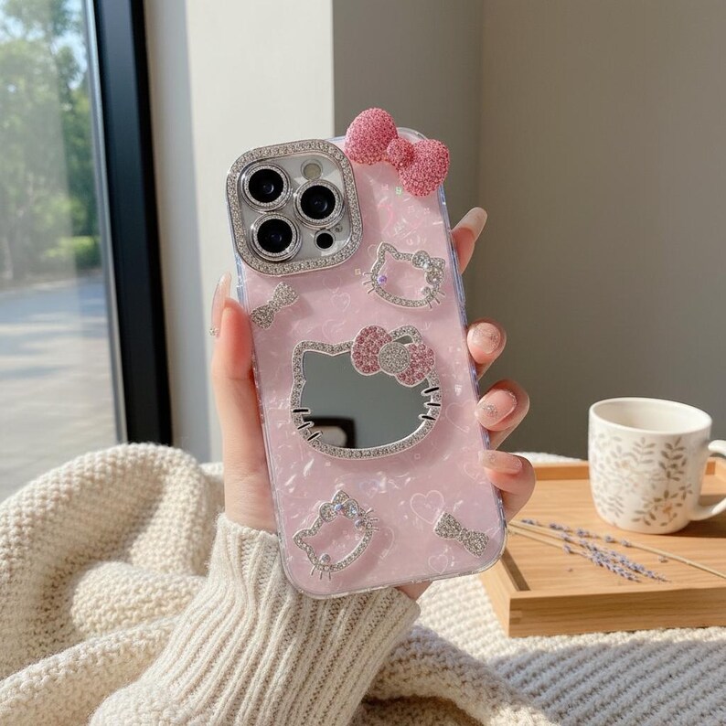 K&ouml;nnte beinhalten: Rosa Handyh&uuml;lle mit Hello Kitty-Design, Spiegel, Schleifendetails und Strassverzierungen. Die H&uuml;lle hat ein klares, sch&uuml;tzendes Design mit einer dekorativen Schleife oben. Die Kameralinse ist von Strasssteinen umgeben.