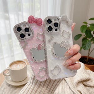 K&ouml;nnte beinhalten: Zwei dekorative Handyh&uuml;llen, eine rosa und eine wei&szlig;e, verziert mit funkelnden Strasssteinen. Jede H&uuml;lle hat eine Schleife und ein Hello Kitty-Design. Die H&uuml;llen sind f&uuml;r ein Smartphone konzipiert und haben einen Objektivschutz.