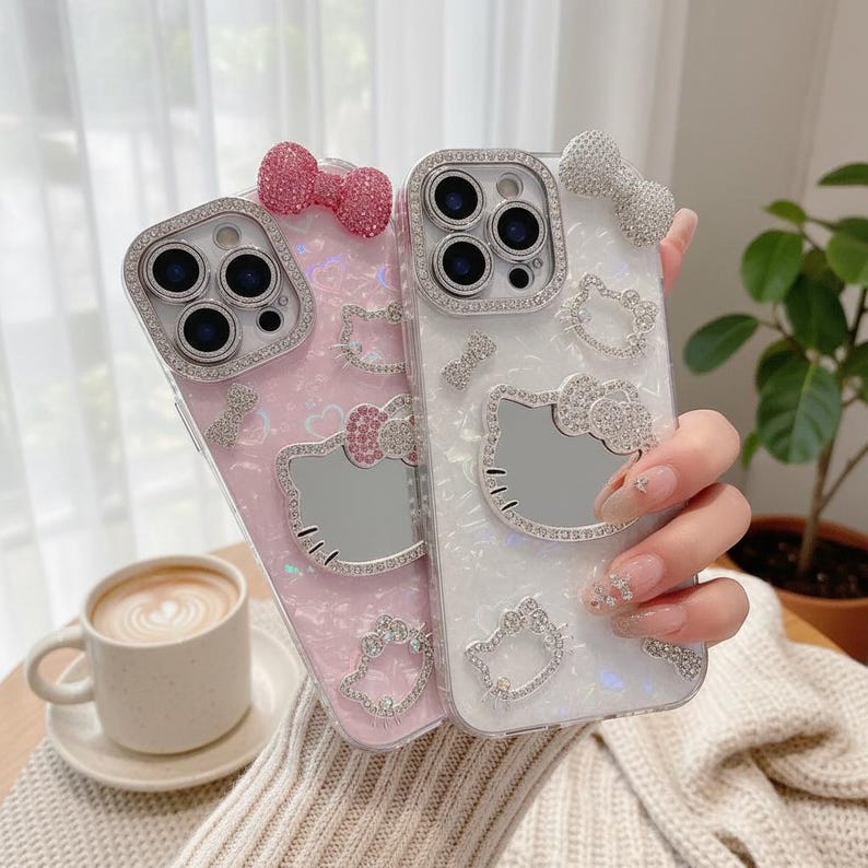 K&ouml;nnte beinhalten: Zwei dekorative Handyh&uuml;llen, eine rosa und eine wei&szlig;e, verziert mit funkelnden Strasssteinen. Jede H&uuml;lle hat eine Schleife und ein Hello Kitty-Design. Die H&uuml;llen sind f&uuml;r ein Smartphone konzipiert und haben einen Objektivschutz.