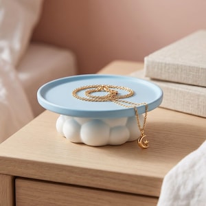 May include: A light blue circular ceramic tray with a raised edge, supported by a white cloud-shaped base. A delicate gold chain necklace with a small crescent moon and star pendant rests on the tray, with the pendant hanging over the edge.