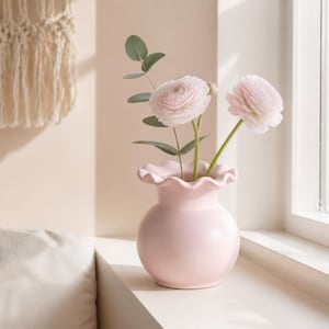 May include: A light pink ceramic vase with a rounded body and a ruffled, scalloped rim. The vase contains two pale pink ranunculus flowers and a single stem of green eucalyptus leaves, creating a soft, balanced floral arrangement in a simple vessel.