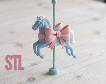 Op de afbeelding: Een lichtblauw carrouselpaard-figuurtje met witte manen en staart, versierd met een grote roze strik. Het paard is gemonteerd op een lichtblauwe paal met een ronde voet. De letters STL staan in het roze aan de linkerkant afgebeeld.
