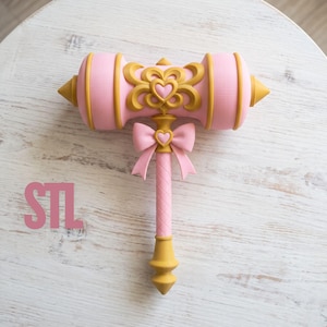 Op de afbeelding: Een roze en gouden speelgoedhamer met een centraal hartmotief op de kop, een roze strik met een klein hartje bovenaan de steel, en de letters STL in het roze gedrukt aan de linkerkant van het object.