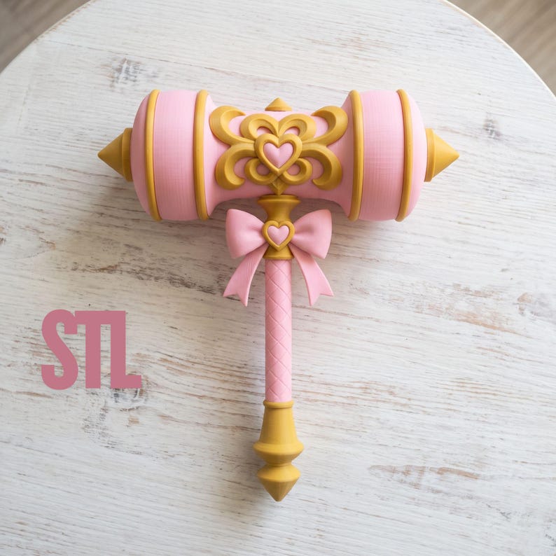 Op de afbeelding: Een roze en gouden speelgoedhamer met een centraal hartmotief op de kop, een roze strik met een klein hartje bovenaan de steel, en de letters STL in het roze gedrukt aan de linkerkant van het object.