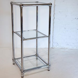 May include: A chrome-finished, three-tiered display shelf with clear glass shelves. The rectangular frame has rounded corners and a polished, reflective surface. The shelf is designed for displaying items and has a minimalist aesthetic.