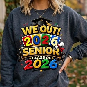 May include: Dark gray sweatshirt with the text "WE OUT! 2026 SENIOR CLASS OF 2026" in yellow, red, green, and blue. The design includes a graduation cap, diploma, and stars. A gold necklace and watch are visible.