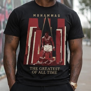 May include: Black t-shirt featuring a graphic of Muhammad Ali in a boxing pose, with the text "MUHAMMAD" above and "THE GREATEST OF ALL TIME" below. The design includes red and white accents.