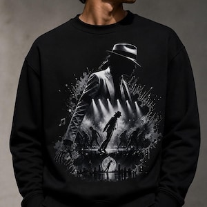 May include: Black sweatshirt featuring a grayscale illustration of a performer in a fedora and suit, dancing on a stage. The design includes musical notes and a cityscape silhouette, creating a dynamic and artistic representation of a music icon.