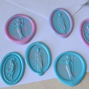 May include: A collection of oval wax seals in shades of light blue and pink, each featuring a raised relief of a classical figure. The seals are arranged on a white envelope alongside a glass dip pen, a wooden wax stamp, and colored wax sticks.