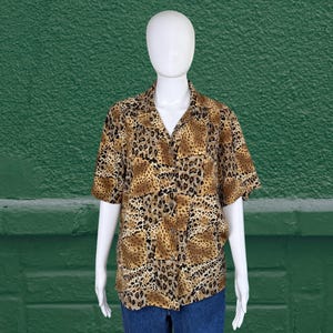 May include: A short-sleeved button-down shirt with a leopard print pattern in shades of brown, tan, and black. The shirt is displayed on a mannequin with blue jeans. The shirt has a collared neckline and two chest pockets.