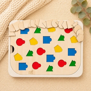 Könnte beinhalten: Ein rechteckiges Holzpuzzle-Brett mit verschiedenen Aussparungen in geometrischen Formen. Das Brett enthält rote, blaue, gelbe und grüne Einsätze, wobei zusätzliche unbemalte Holzpuzzleteile am oberen Rand liegen. Das Laser Lux Logo ist sichtbar.