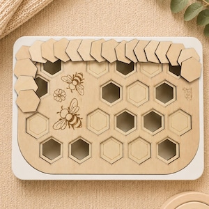 Può includere: Una tavola puzzle rettangolare in legno con cornice bianca caratterizzata da un motivo a griglia a nido d'ape. La tavola include illustrazioni incise di due api e un fiore, con diversi pezzi esagonali in legno sparsi sul bordo.