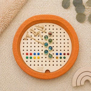 Könnte beinhalten: Ein Solitärspiel aus Holz mit einem orangefarbenen Rand. Das Spielbrett hat eine helle Holzmitte mit gleichmäßig verteilten Löchern. Holzstifte und bunte Murmeln sind auf dem Brett platziert. Der Markenname "LaserLux" ist auf dem Brett aufgedruckt.