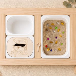 Op de afbeelding: Een houten sensorisch dienblad met drie witte compartimenten. Eén compartiment bevat zand en kleurrijke edelstenen. Een houten lepel is inbegrepen. Een houten schatkistontwerp staat op een houten inzetstuk.
