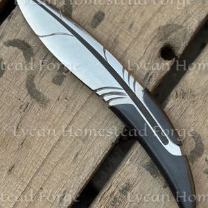Puede incluir: Un cuchillo de metal con una hoja en forma de pluma, con un acabado met&aacute;lico plateado con detalles grabados y un mango negro s&oacute;lido. El texto Lycan Homestead Forge es visible en la imagen, superponi&eacute;ndose al cuchillo y a la superficie.