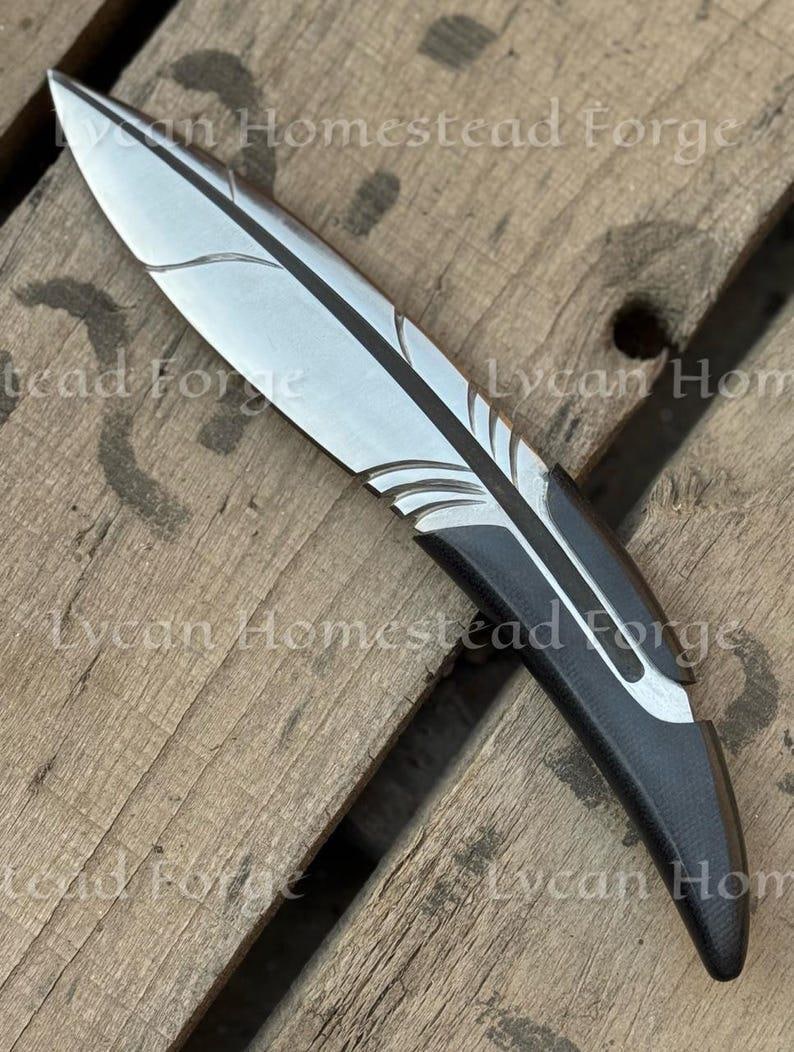 Puede incluir: Un cuchillo de metal con una hoja en forma de pluma, con un acabado met&aacute;lico plateado con detalles grabados y un mango negro s&oacute;lido. El texto Lycan Homestead Forge es visible en la imagen, superponi&eacute;ndose al cuchillo y a la superficie.