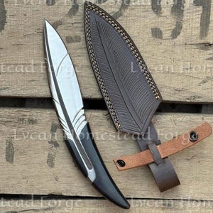 Puede incluir: Un cuchillo de metal plateado con una hoja en forma de pluma y un mango negro, acompa&ntilde;ado de una funda de cuero marr&oacute;n oscuro con un patr&oacute;n de pluma en relieve a juego y una correa de cuero marr&oacute;n claro con dos botones de presi&oacute;n met&aacute;licos.