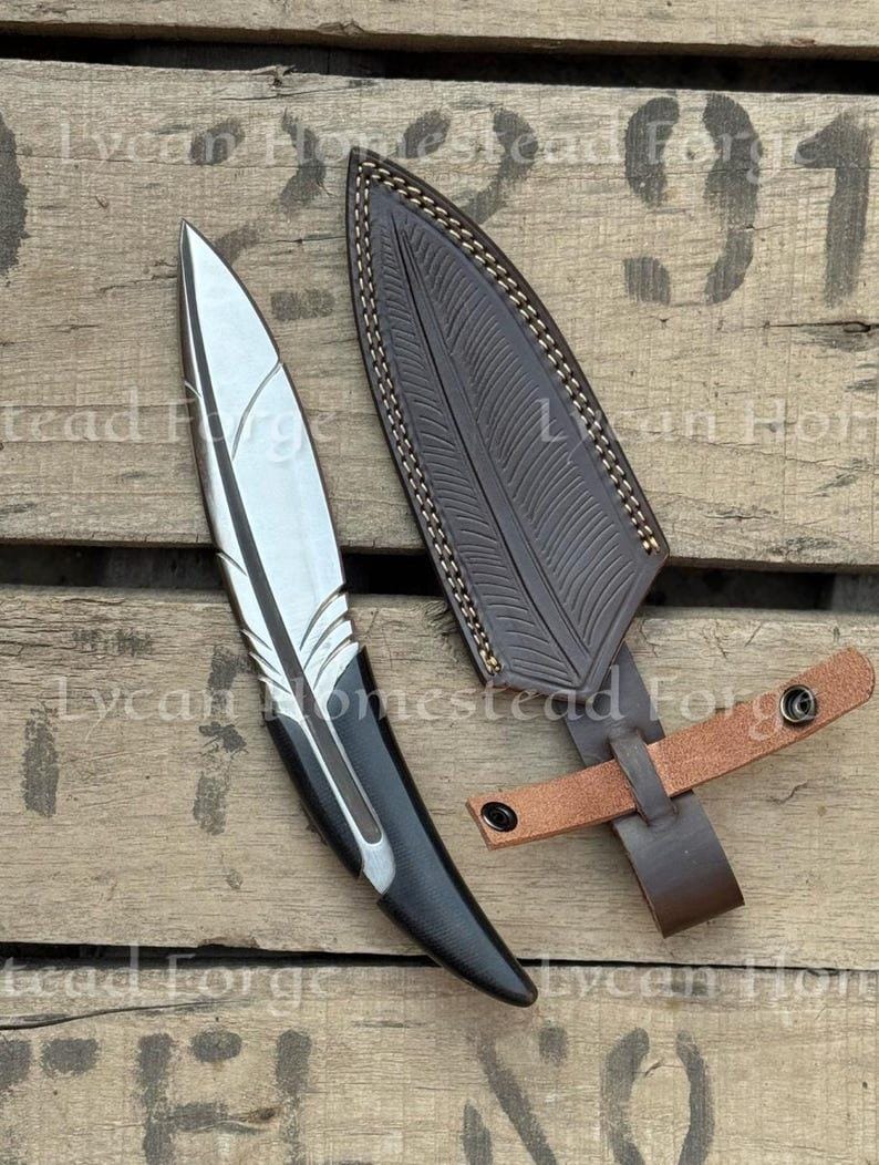 Puede incluir: Un cuchillo de metal plateado con una hoja en forma de pluma y un mango negro, acompa&ntilde;ado de una funda de cuero marr&oacute;n oscuro con un patr&oacute;n de pluma en relieve a juego y una correa de cuero marr&oacute;n claro con dos botones de presi&oacute;n met&aacute;licos.
