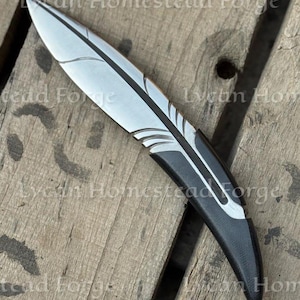 Puede incluir: Un cuchillo de metal con forma de pluma, con una hoja plateada que presenta l&iacute;neas grabadas y un mango negro s&oacute;lido. El texto Lycan Homestead Forge es visible en la imagen. El cuchillo tiene un dise&ntilde;o curvo con una punta afilada y acabado liso.