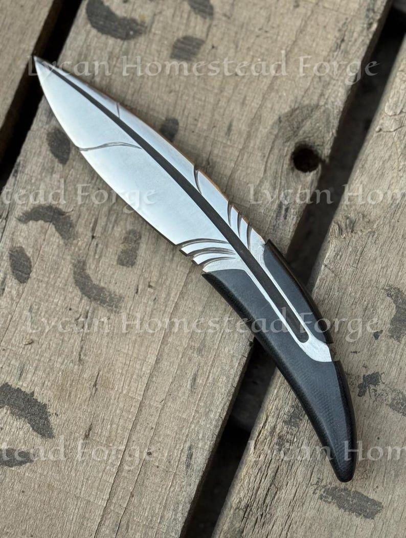 Puede incluir: Un cuchillo de metal con forma de pluma, con una hoja plateada que presenta l&iacute;neas grabadas y un mango negro s&oacute;lido. El texto Lycan Homestead Forge es visible en la imagen. El cuchillo tiene un dise&ntilde;o curvo con una punta afilada y acabado liso.