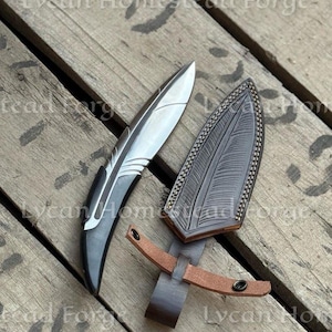 Puede incluir: Un cuchillo de hoja fija con una hoja de metal plateado y un mango oscuro y curvo, acompa&ntilde;ado de una funda de cuero marr&oacute;n oscuro. La funda presenta un patr&oacute;n en relieve detallado en forma de pluma y una correa de cuero tostado con broche.