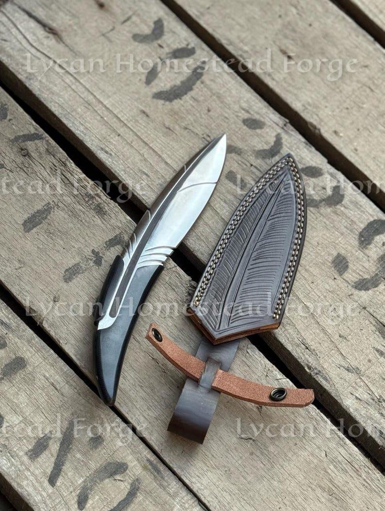 Puede incluir: Un cuchillo de hoja fija con una hoja de metal plateado y un mango oscuro y curvo, acompa&ntilde;ado de una funda de cuero marr&oacute;n oscuro. La funda presenta un patr&oacute;n en relieve detallado en forma de pluma y una correa de cuero tostado con broche.