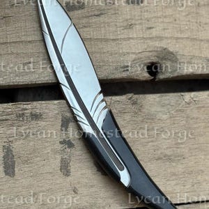 Puede incluir: Un abrecartas de metal con forma de pluma, punta plateada y mango negro. La hoja presenta l&iacute;neas grabadas que imitan la estructura de una pluma. El texto Lycan Homestead Forge es visible a lo largo de toda la imagen.
