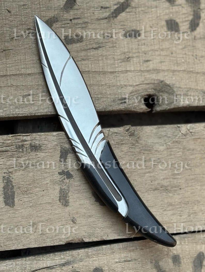 Puede incluir: Un abrecartas de metal con forma de pluma, punta plateada y mango negro. La hoja presenta l&iacute;neas grabadas que imitan la estructura de una pluma. El texto Lycan Homestead Forge es visible a lo largo de toda la imagen.