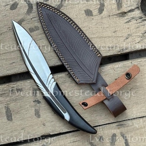 Puede incluir: Un cuchillo de hoja fija con una hoja de acero plateada en forma de pluma y un mango negro, acompa&ntilde;ado de una funda de cuero marr&oacute;n oscuro con un patr&oacute;n de pluma estampado, costuras claras y una correa de cuero beige con dos broches met&aacute;licos.