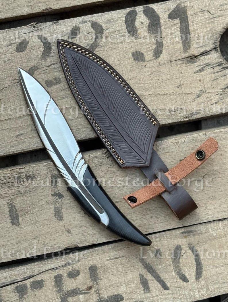 Puede incluir: Un cuchillo de hoja fija con una hoja de acero plateada en forma de pluma y un mango negro, acompa&ntilde;ado de una funda de cuero marr&oacute;n oscuro con un patr&oacute;n de pluma estampado, costuras claras y una correa de cuero beige con dos broches met&aacute;licos.