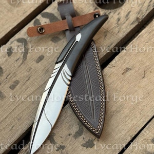 Puede incluir: Un cuchillo de hoja fija con una hoja de metal plateado en forma de pluma, con un mango gris oscuro y una funda de cuero marr&oacute;n oscuro a juego con detalles de plumas en relieve y un cierre de correa con bot&oacute;n a presi&oacute;n.