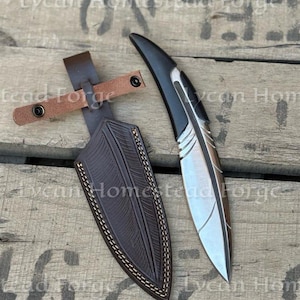 Puede incluir: Un cuchillo de hoja fija con un mango curvo en forma de pluma y una hoja met&aacute;lica, acompa&ntilde;ado de una funda de cuero marr&oacute;n oscuro con un patr&oacute;n de pluma grabado y una trabilla de cuero marr&oacute;n claro con dos broches de presi&oacute;n negros.