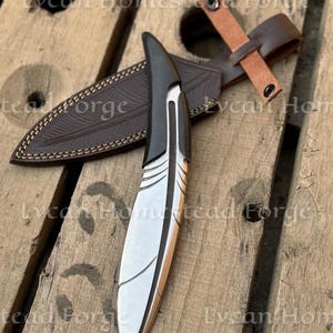 Puede incluir: Un cuchillo de metal plateado con mango negro en forma de pluma y l&iacute;neas grabadas en la hoja. Se acompa&ntilde;a de una funda de cuero marr&oacute;n oscuro con patr&oacute;n de plumas y una correa de cuero beige con un bot&oacute;n de presi&oacute;n met&aacute;lico.