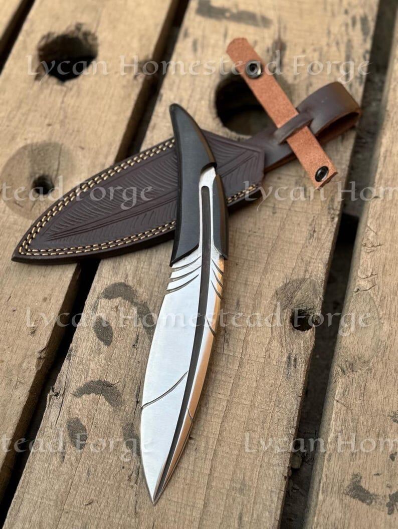 Puede incluir: Un cuchillo de metal plateado con mango negro en forma de pluma y l&iacute;neas grabadas en la hoja. Se acompa&ntilde;a de una funda de cuero marr&oacute;n oscuro con patr&oacute;n de plumas y una correa de cuero beige con un bot&oacute;n de presi&oacute;n met&aacute;lico.