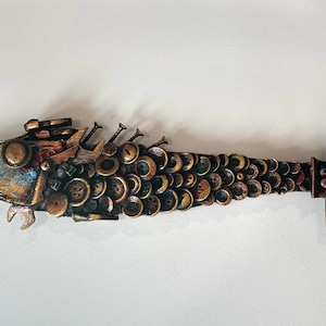 Puede incluir: Escultura de pez artesanal de técnica mixta. El pez presenta una cabeza con detalles metálicos, un cuerpo compuesto por numerosos botones y una cola con un marco rectangular. Incorpora clavos y otros elementos metálicos, creando una apariencia texturizada única.