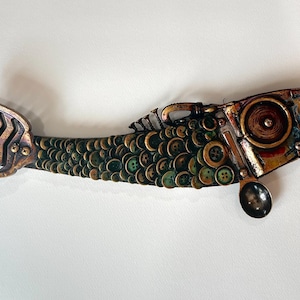 Puede incluir: Una escultura de pez artesanal hecha con materiales reciclados. El cuerpo está cubierto de botones verdes y marrones, con una cola y cabeza de metal. Una cuchara está incorporada en el diseño, agregando un toque único a esta pieza decorativa.