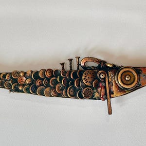 Könnte beinhalten: Eine handgefertigte Fischskulptur im Steampunk-Stil. Der Körper besteht aus Knöpfen, Zahnrädern und Metallteilen mit Kupfer- und Bronze-Finish. Schwanz und Kopf sind mit einem Farbverlauf aus Orange und Braun bemalt.