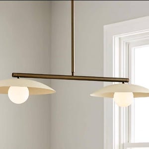Puede incluir: Una lámpara colgante moderna de dos luces con acabado bronce. La lámpara presenta dos pantallas en forma de platillo de color blanquecino y bombillas esféricas de vidrio esmerilado. El accesorio se suspende de una barra y una barra horizontal.