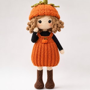 May include: A handmade crochet doll featuring curly brown hair, wearing a pumpkin-shaped hat, a black long-sleeved shirt, and an orange pinafore dress with a small pumpkin detail. The doll has dark eyes, rosy cheeks, and brown boots.