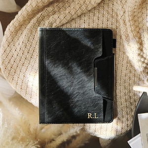 May include: A black leather-textured notebook cover featuring a vertical pocket with a silver-toned metal accent and the gold-embossed initials R.L. in the bottom right corner. The rectangular cover has visible stitching along the edges.
